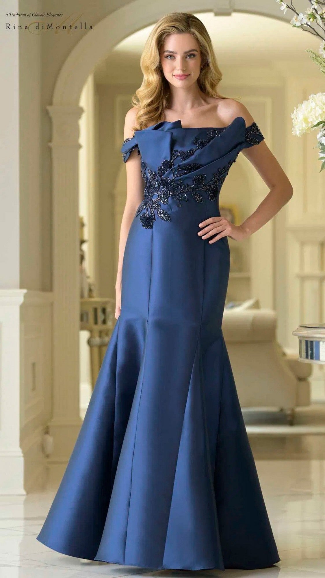 A woman indoors wears the Rina Di Montella RD3408, a navy long formal dress with an off-the-shoulder neckline, delicate floral embellishments, and a floor-length mermaid silhouette.